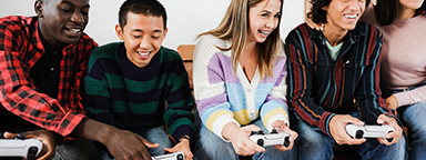 Fünf junge Gamer sitzen zu Hause auf einer Couch, vier halten PS5-Controller in der Hand und spielen zusammen.