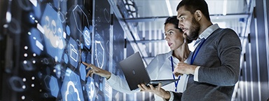 Especialista informático sosteniendo un portátil mientras habla con un técnico en la sala de servidores de un centro de datos, con iconos de nube y escudo en los bastidores de servidores