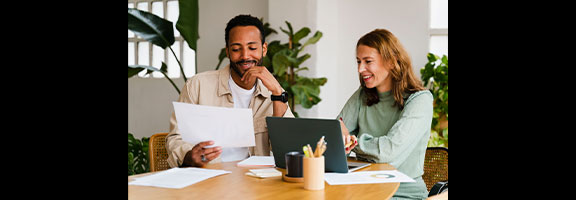 Two coworkers still at a table, one is holding a paper, the other is on a laptop