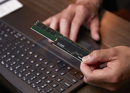 Der Unterschied zwischen Arbeitsspeicher und Storage (Laufwerks-Speicher) Nahaufnahme einer Hand, die eine Kingston KC3000 SSD und ein DDR5-Speichermodul hält, dahinter ein Laptop auf einem Schreibtisch